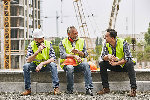 tres hombres con casco y chaleco sentados conversando en una construcción